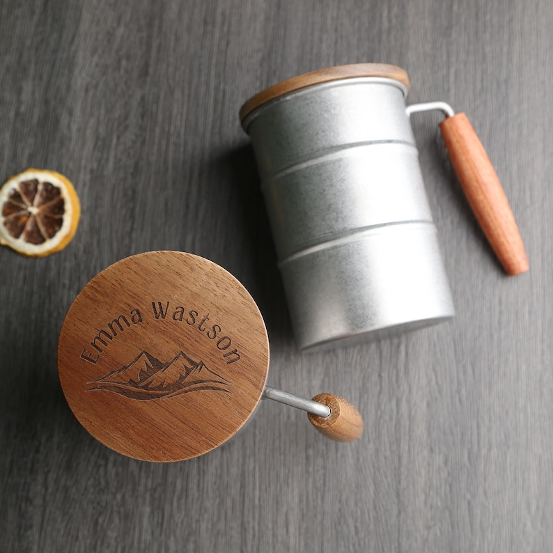May include: A wooden lid with the text "Emma Watson" and a mountain range design. The lid is attached to a silver measuring cup with a wooden handle.