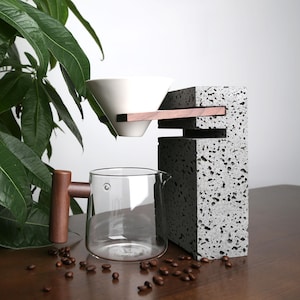 Op de afbeelding: Een grijze betonnen koffiezetapparaat met een houten handvat en een wit keramisch filter. Het koffiezetapparaat staat op een houten tafel met koffiebonen eromheen.
