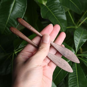 May include: Two wooden butter knives with a pointed tip. The knives are made of dark brown wood and have the word "ธรรมดา" engraved on one of the knives.
