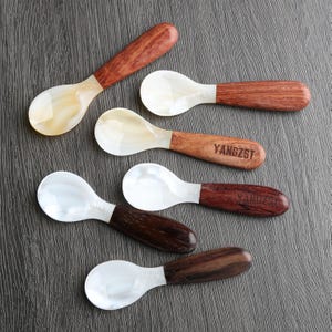 May include: Five spoons with white mother of pearl bowls and wooden handles. The handles are made of different types of wood, including dark brown, light brown, and reddish brown. The spoons are arranged in a semi-circle on a dark gray surface. The spoons are engraved with the word "YANGZST".