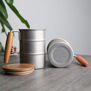 May include: Two silver stainless steel mugs with wooden handles and lids. The mugs are stacked on top of each other. The mugs are on a gray surface.