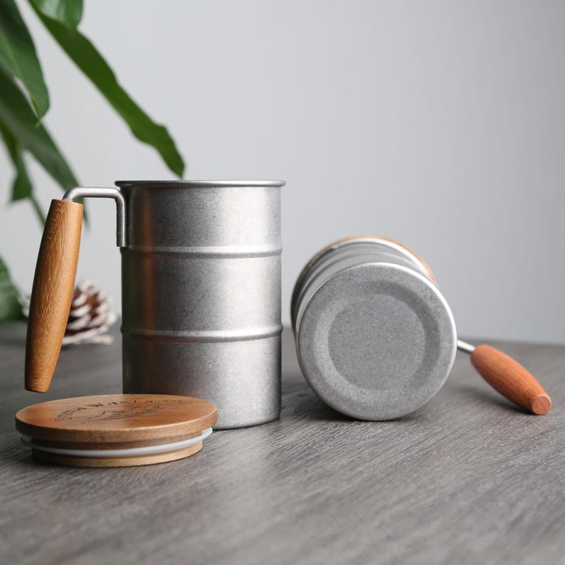 May include: Two silver stainless steel mugs with wooden handles and lids. The mugs are stacked on top of each other. The mugs are on a gray surface.