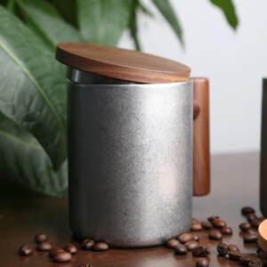 Pode incluir: Uma caneca de cer&acirc;mica prateada com tampa e al&ccedil;a de madeira. A caneca est&aacute; sobre uma superf&iacute;cie de madeira com gr&atilde;os de caf&eacute; espalhados ao redor.
