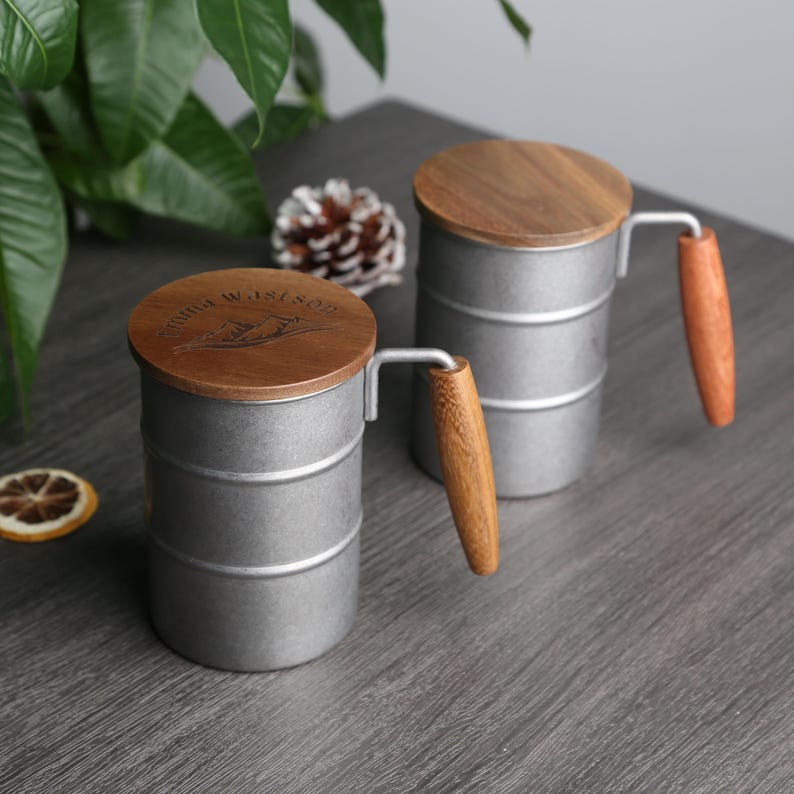 May include: Two silver metal mugs with wooden lids and handles. The lids have the words "Mountain Waistline" engraved on them.