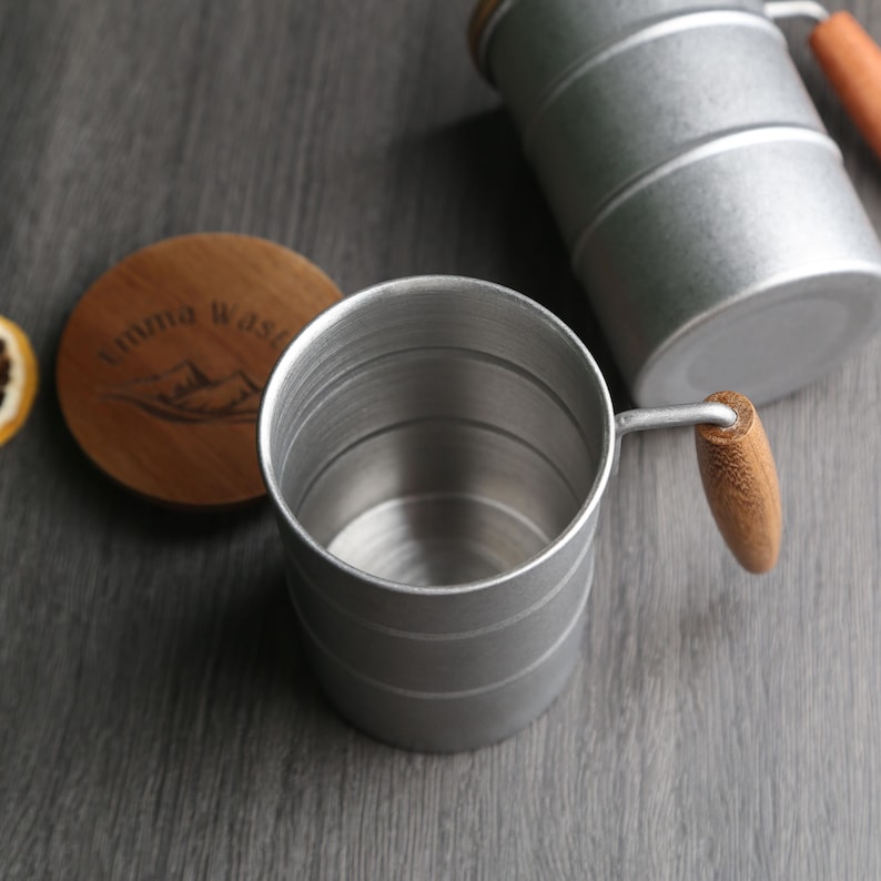 May include: A silver collapsible camping mug with a wooden handle. The mug is made of metal and has a smooth, polished finish. The handle is made of natural wood and has a comfortable grip.