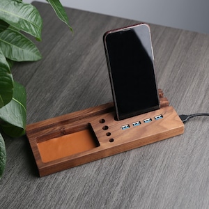 May include: A wooden phone stand with a built-in charging station. The stand has a leather-lined compartment for holding small items and multiple USB ports for charging devices. The stand is made of dark wood and has a sleek, modern design.