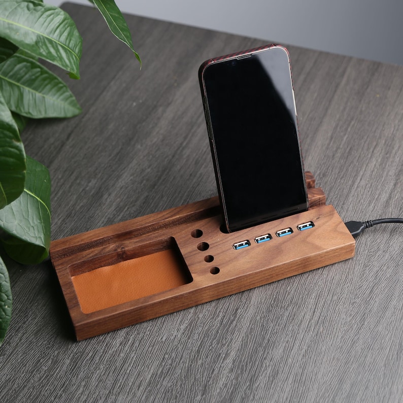 May include: A wooden phone stand with a built-in charging station. The stand has a leather-lined compartment for holding small items and multiple USB ports for charging devices. The stand is made of dark wood and has a sleek, modern design.