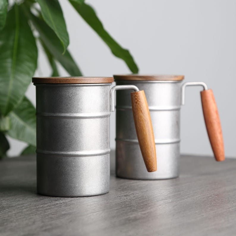 May include: Two silver metal mugs with wooden lids and handles. The mugs are on a wooden surface.
