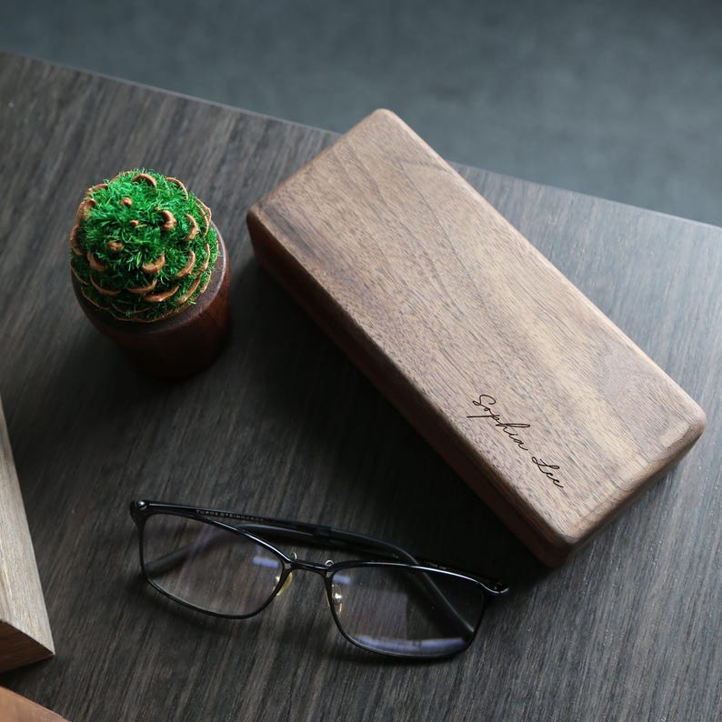 May include: A wooden rectangular box with a dark brown finish. The box has a personalized inscription that reads "Sophia Lee". The box is sitting on a dark wood surface with a small green plant in a pot next to it. A pair of black eyeglasses is also on the surface.
