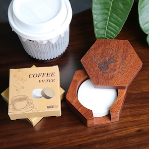 May include: A wooden, hexagonal coffee filter holder with a cat design, next to a box of coffee filters and a white plastic container. The box reads "COFFEE FILTER" and "100 pcs". The wooden holder is open, revealing white filters.