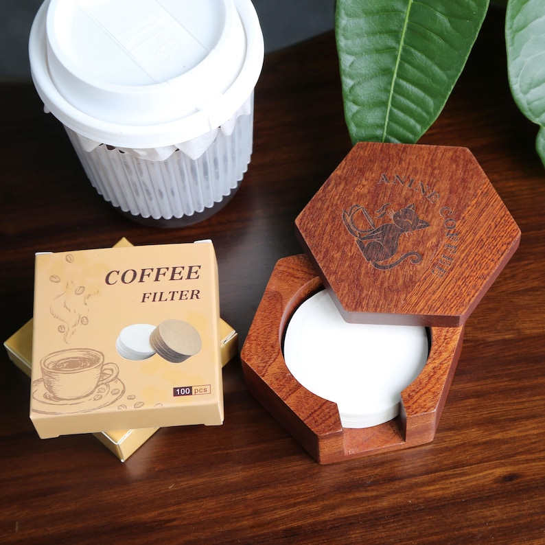 May include: A wooden, hexagonal coffee filter holder with a cat design, next to a box of coffee filters and a white plastic container. The box reads "COFFEE FILTER" and "100 pcs". The wooden holder is open, revealing white filters.