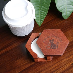 May include: A wooden, hexagonal coffee filter holder with a hinged lid. The lid is engraved with a cat and the words "ANIME COFFEE". A white paper filter is visible inside. A white coffee cup with a lid is in the background.