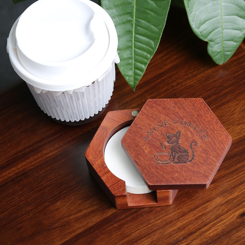 May include: A wooden, hexagonal coffee filter holder with a hinged lid. The lid is engraved with a cat and the words "ANIME COFFEE". A white paper filter is visible inside. A white coffee cup with a lid is in the background.