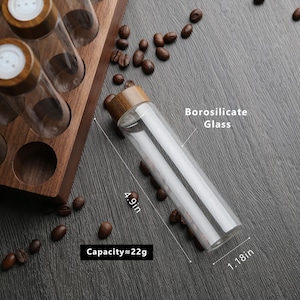 Puede incluir: Una botella de vidrio transparente con una tapa de madera, que mide 12,4 cm de alto y 3 cm de di&aacute;metro. La botella est&aacute; etiquetada como "Vidrio de borosilicato" y tiene una capacidad de 22 gramos. La botella est&aacute; sobre una superficie oscura con granos de caf&eacute; esparcidos a su alrededor.