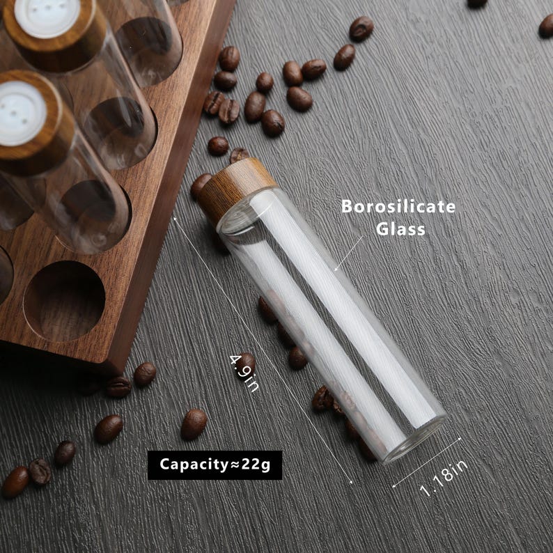 Puede incluir: Una botella de vidrio transparente con una tapa de madera, que mide 12,4 cm de alto y 3 cm de di&aacute;metro. La botella est&aacute; etiquetada como "Vidrio de borosilicato" y tiene una capacidad de 22 gramos. La botella est&aacute; sobre una superficie oscura con granos de caf&eacute; esparcidos a su alrededor.