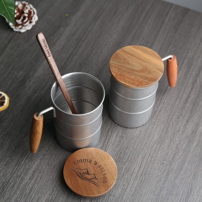 May include: Two silver camping mugs with wooden lids and handles. One mug has a wooden spoon with the text "Ann Rose" engraved on it. The other mug has a wooden lid with the text "Emma Watson" engraved on it.