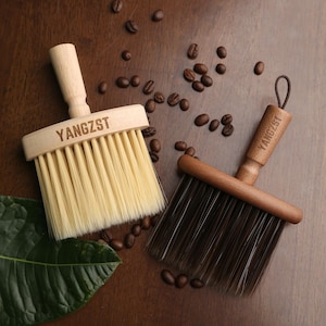 Peut inclure: Deux brosses à poussière à manche en bois avec le mot "YANGZST" imprimé sur les manches. La brosse de gauche a des poils brun clair et la brosse de droite a des poils brun foncé. Les deux brosses sont sur une surface en bois brun avec des grains de café éparpillés autour d'elles.