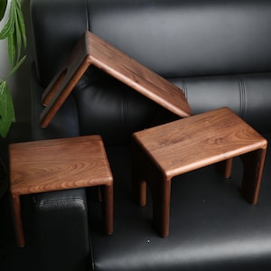 May include: Three wooden stools in varying sizes. The stools are made of a dark brown wood with a smooth finish. One stool is rectangular, another is a small square, and the third is a long, angled piece. The stools are on a black surface.