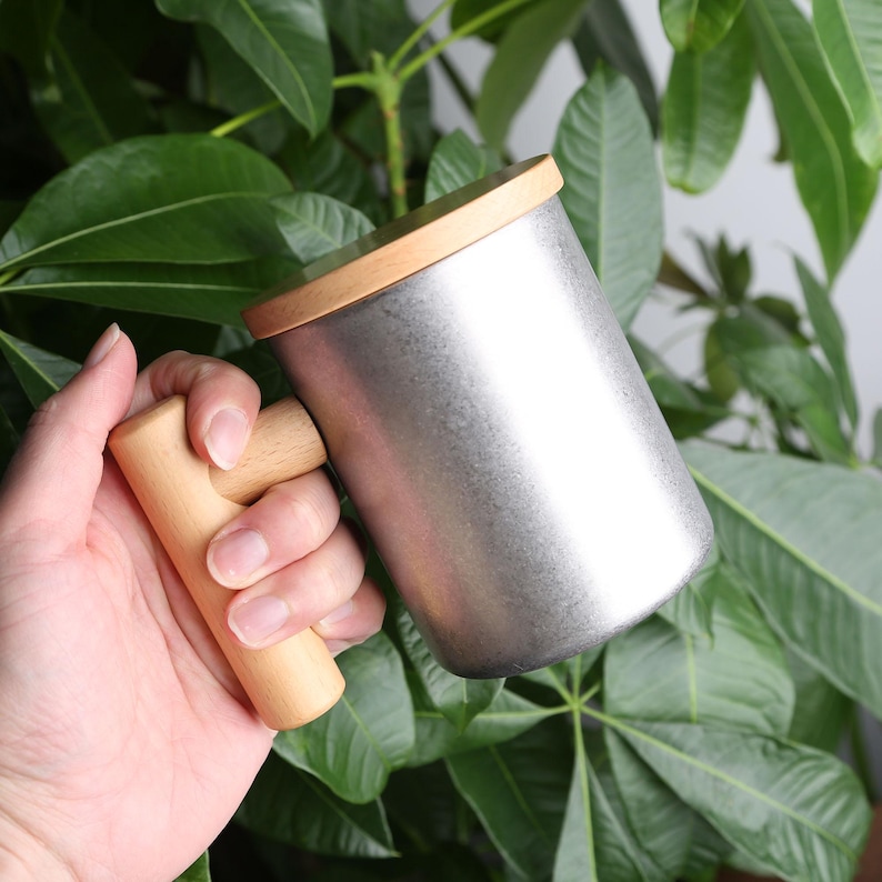 Pode incluir: Uma caneca de metal prateada com uma al&ccedil;a de madeira e uma tampa de madeira. A caneca est&aacute; sobre uma planta verde com folhas.