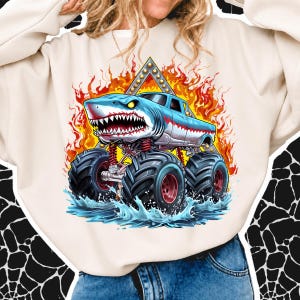 May include: A cream-colored crewneck sweatshirt featuring a graphic print of a blue monster truck designed to look like a shark with sharp teeth, large black tires, and red suspension, set against a background of orange flames and water.