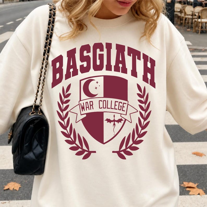 May include: A cream-colored crewneck sweatshirt featuring a maroon graphic print that reads BASGIATH WAR COLLEGE. The design includes a shield emblem with a crescent moon, stars, and a dragon silhouette, flanked by two laurel leaf branches.