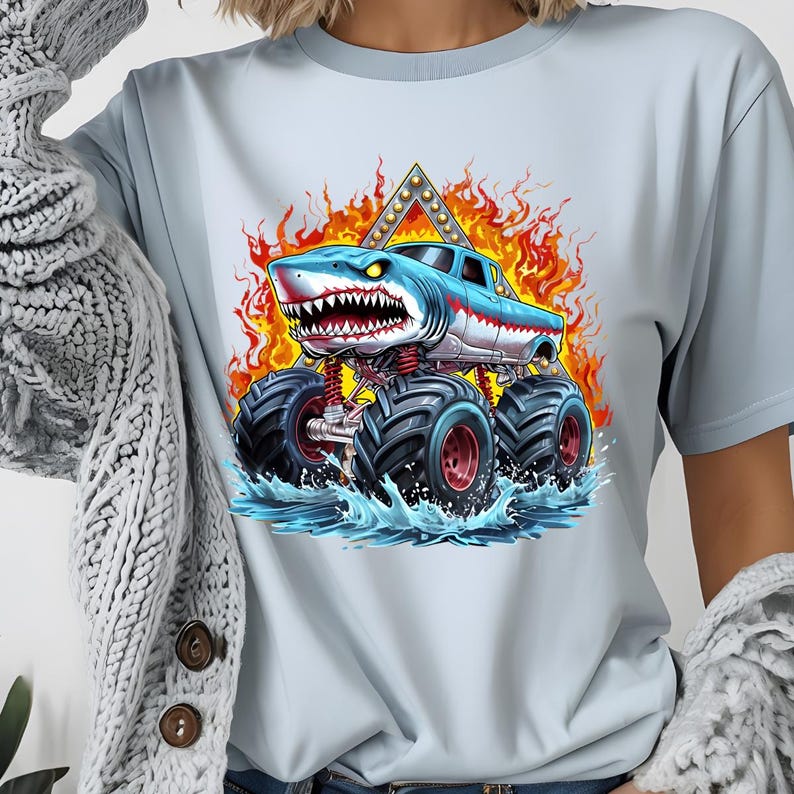 May include: A light blue t-shirt with a graphic of a blue and white monster truck with a shark head. The truck is driving through water and flames.
