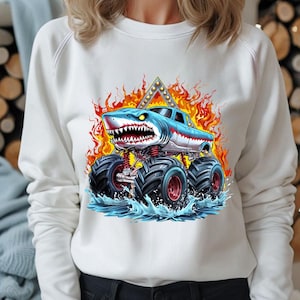 May include: White sweatshirt with a graphic of a blue and white monster truck with a shark head. The truck is driving through water and flames.