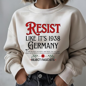 May include: White sweatshirt with the text "RESIST LIKE IT'S 1938 GERMANY" in red and black lettering. The text "IT TOOK THE NAZIS ONE MONTH, THREE WEEKS, TWO DAYS, EIGHT HOURS AND 40 MINUTES TO DISMANTLE A CONSTITUTIONAL REPUBLIC" is printed in black below the main text. A red flower with green leaves is printed below the text. The text "REJECT FASCISTS" is printed in black below the flower.