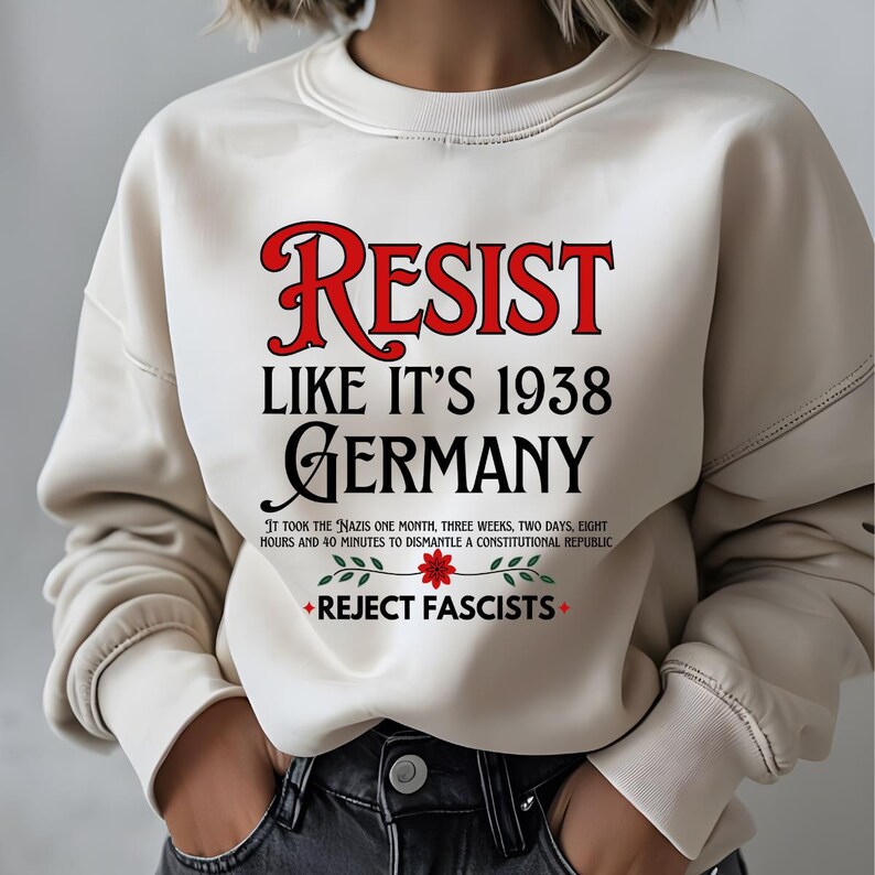 May include: White sweatshirt with the text "RESIST LIKE IT'S 1938 GERMANY" in red and black lettering. The text "IT TOOK THE NAZIS ONE MONTH, THREE WEEKS, TWO DAYS, EIGHT HOURS AND 40 MINUTES TO DISMANTLE A CONSTITUTIONAL REPUBLIC" is printed in black below the main text. A red flower with green leaves is printed below the text. The text "REJECT FASCISTS" is printed in black below the flower.