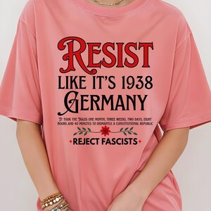 May include: Pink t-shirt with the text "RESIST LIKE IT'S 1938 GERMANY" in black and red. The text "IT TOOK THE NAZIS ONE MONTH, THREE WEEKS, TWO DAYS, EIGHT HOURS AND 40 MINUTES TO DISMANTLE A CONSTITUTIONAL REPUBLIC" is printed in black below the main text. A red flower with green leaves is printed below the text. The text "REJECT FASCISTS" is printed in black below the flower.