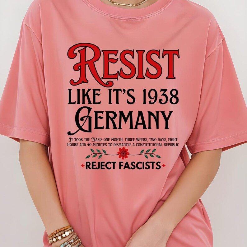 May include: Pink t-shirt with the text "RESIST LIKE IT'S 1938 GERMANY" in black and red. The text "IT TOOK THE NAZIS ONE MONTH, THREE WEEKS, TWO DAYS, EIGHT HOURS AND 40 MINUTES TO DISMANTLE A CONSTITUTIONAL REPUBLIC" is printed in black below the main text. A red flower with green leaves is printed below the text. The text "REJECT FASCISTS" is printed in black below the flower.