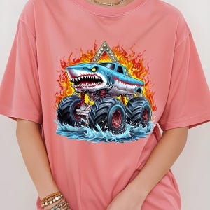 May include: A pink t-shirt with a graphic of a blue monster truck with shark teeth, flames, and water splashing around it.
