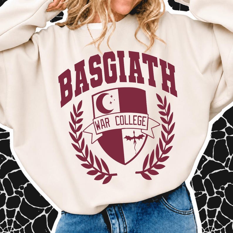 May include: A cream-colored crewneck sweatshirt featuring a maroon graphic on the chest. The design includes the text BASGIATH WAR COLLEGE in an arched font above a shield emblem containing a crescent moon, stars, and a dragon silhouette.