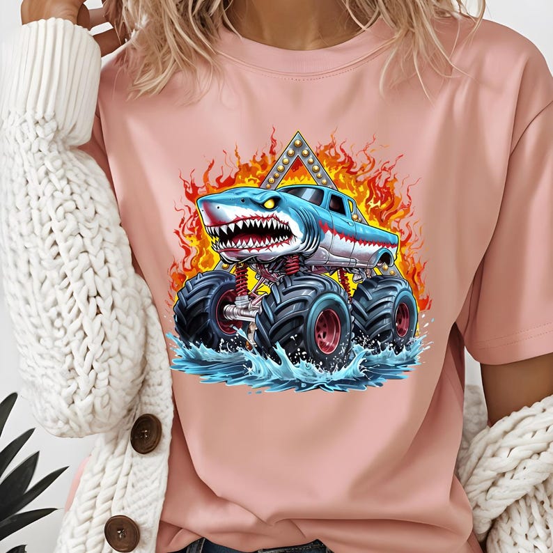 May include: Pink t-shirt with a graphic of a blue shark monster truck with flames and water splashes.