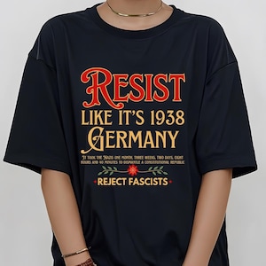 May include: Black t-shirt with the text "RESIST LIKE IT'S 1938 GERMANY" in red and gold lettering. Below the text is a floral design with the text "REJECT FASCISTS" in red.