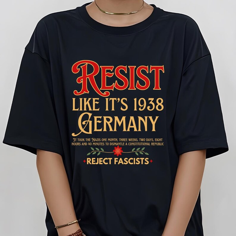 May include: Black t-shirt with the text "RESIST LIKE IT'S 1938 GERMANY" in red and gold lettering. Below the text is a floral design with the text "REJECT FASCISTS" in red.