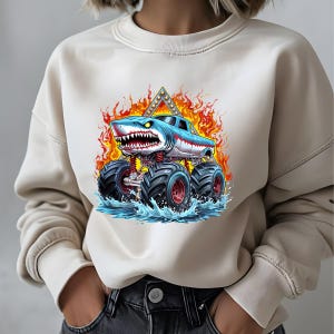 May include: A white sweatshirt with a graphic of a blue monster truck with a shark head, flames, and water.