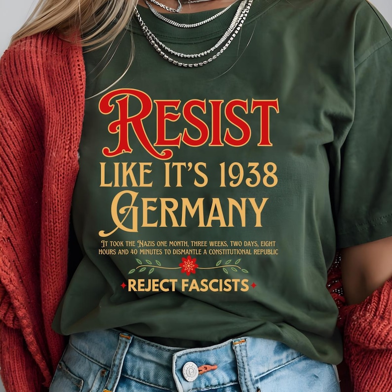 May include: Green t-shirt with the text "RESIST LIKE IT'S 1938 GERMANY" in red and yellow lettering. The text "IT TOOK THE NAZIS ONE MONTH, THREE WEEKS, TWO DAYS, EIGHT HOURS AND 40 MINUTES TO DISMANTLE A CONSTITUTIONAL REPUBLIC" is printed in black below the main text. The text "REJECT FASCISTS" is printed in white with a red star on either side.