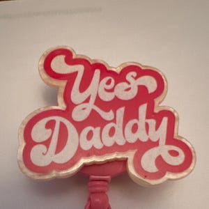 Puede incluir: Encanto de plástico rosa con el texto "Yes Daddy" en letras blancas. El encanto tiene una cuenta en forma de corazón con un diseño plateado y acentos rosados.
