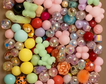 Puede incluir: Una colección de cuentas coloridas de varias formas y tamaños. El surtido incluye cuentas redondas, con forma de Mickey Mouse y con forma de flor en tonos de rosa, amarillo, verde y transparente. Algunas cuentas tienen purpurina o superficies texturizadas.