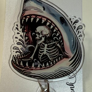 Peut inclure: Un enrouleur de badge avec un motif de requin. La gueule du requin est ouverte, révélant un squelette. Le requin est bleu, blanc et rose, avec des dents noires. Le mot "thank" est écrit en cursive.