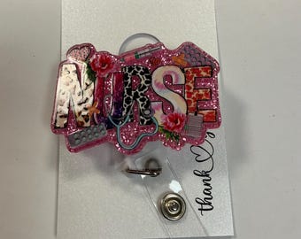 Könnte beinhalten: Rosa glitzernder Badgehalter mit dem Wort "NURSE" in bunten Buchstaben und einem Stethoskop-Design. Der Badgehalter hat ein transparentes Plastikband mit einem Metallclip und zwei Metalldruckknöpfen.