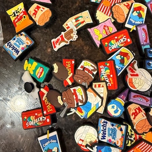 Può includere: Una collezione di ciondoli colorati per scarpe con loghi in miniatura di marchi di cibo e bevande, tra cui KFC, Nutella, Skittles, Capri-Sun, Welch's, Walkers e KitKat. Ogni ciondolo è un piccolo accessorio in plastica piatta e multicolore.
