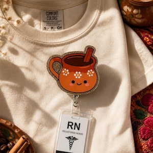 May include: A cream-colored t-shirt with a cute, orange, glittery badge reel featuring a smiling mug of hot chocolate with a cinnamon stick. The badge reel is attached to a clear RN Registered Nurse ID badge.