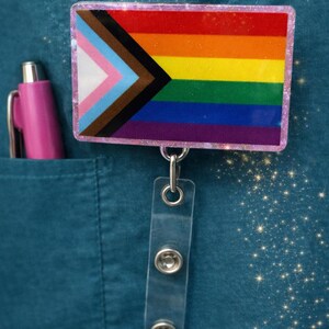 Pode incluir: Um crachá retangular com a bandeira Progress Pride e uma borda rosa brilhante. O crachá está preso a um suporte retrátil de plástico transparente. Uma caneta rosa é visível no bolso de uma peça de roupa verde-azulado.