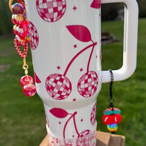 May include: White stainless steel travel mug with a pink and white cherry pattern. The mug has a black chain with a red cherry charm attached to the handle.