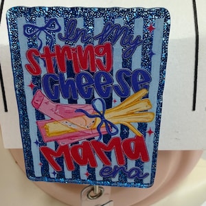 Puede incluir: Un portatarjetas rectangular con acabado de purpurina azul, rayas verticales y el texto In My String Cheese Mama era. El diseño incluye un gráfico de queso en tiras dentro de un envoltorio rosa atado con un lazo azul y pequeñas estrellas rojas.