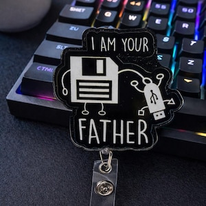 May include: Black and white badge with the text "I AM YOUR FATHER" and a floppy disk and USB drive design. The badge is attached to a clear plastic holder with a metal clip. The background includes a keyboard and a white mug.