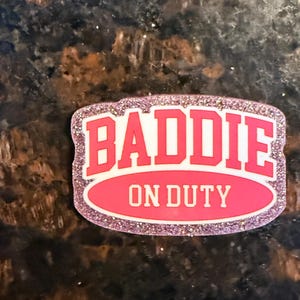 May include: A pink and white rectangular badge with the words "BADIE ON DUTY" in bold letters. The badge has a glittery, pink border. The background is a dark, speckled surface.