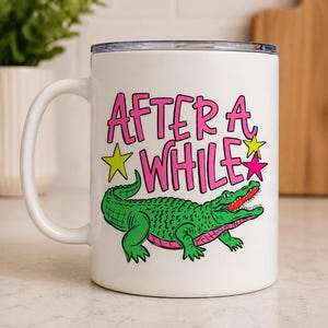 Puede incluir: Taza de cerámica blanca con asa, con una ilustración de un cocodrilo verde con detalles en rosa. La taza tiene el texto "AFTER A WHILE" en rosa con estrellas amarillas y rosas. La taza tiene un borde plateado.